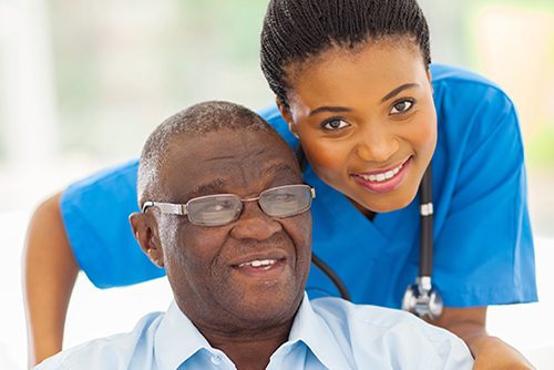 Young nurse in blue scrubs standing behind elderly man in wheelchair both smiling